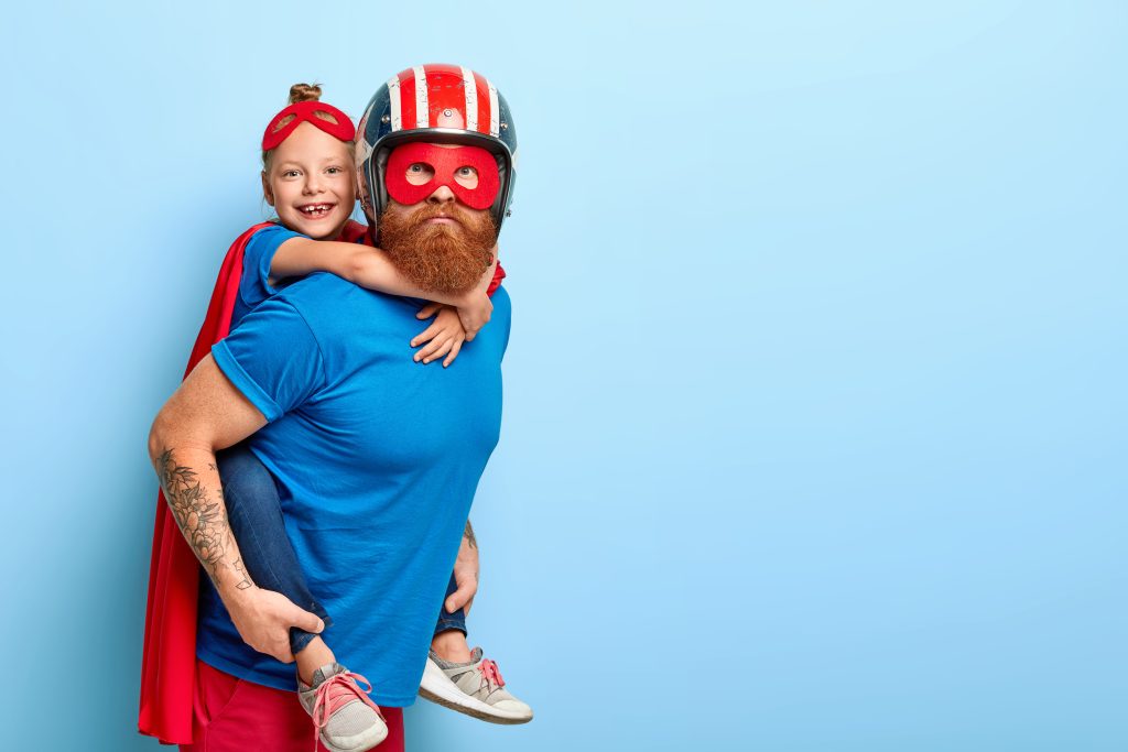 Horizontal shot of caring father gives piggyback to small child, play interesting superhero game, wear special costumes, pose against blue background with free space, have great impact on world